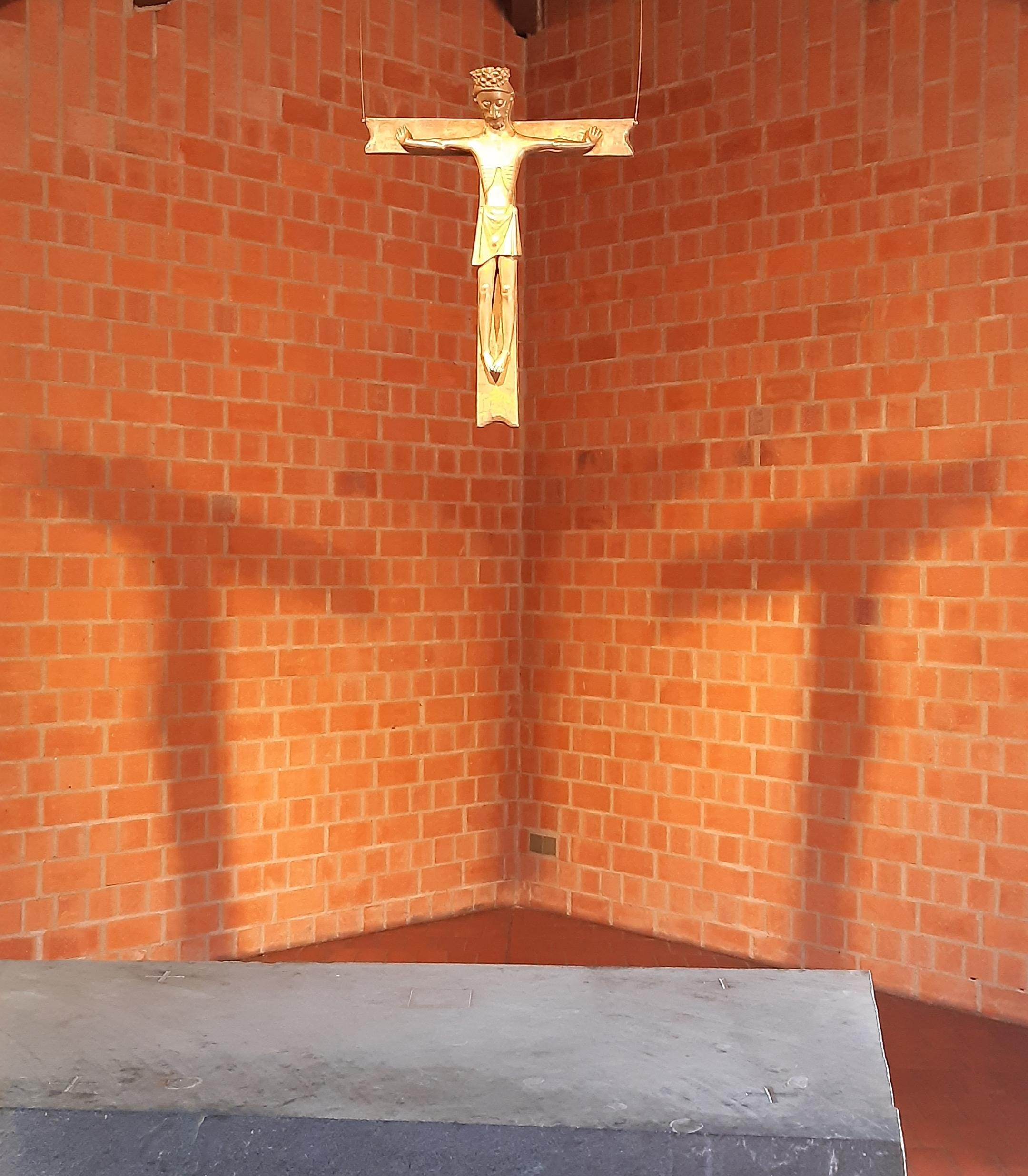 Bronzekreuz mit Christusfigur vor beleuchteter Backsteinwand mit Kreuzschatten