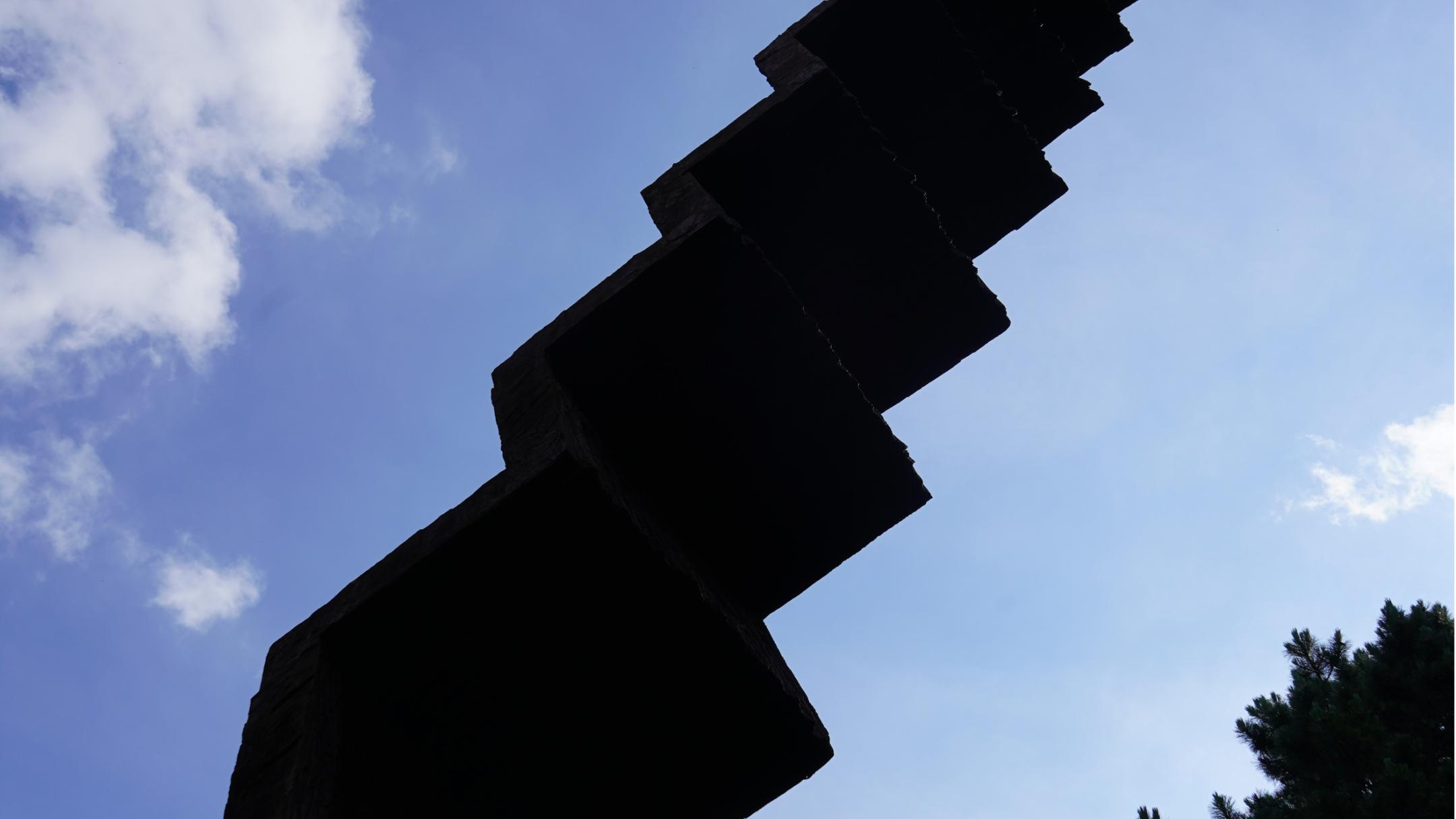 Vertikale, schlanke, angeschrägte Stufenskulptur im Grünen.