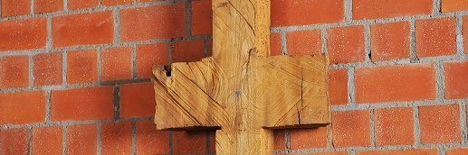Schmale, symmetrische Holzskulptur vor Backsteinwand, erinnert an Kreuz oder archaisches Zeichen.