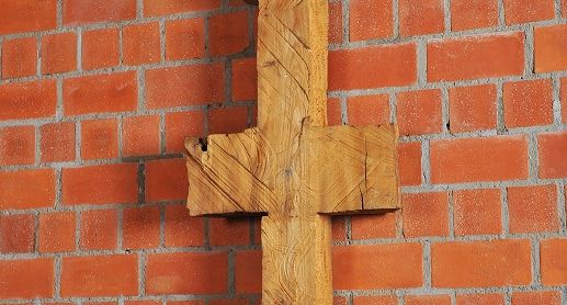 Schmale, symmetrische Holzskulptur vor Backsteinwand, erinnert an Kreuz oder archaisches Zeichen.