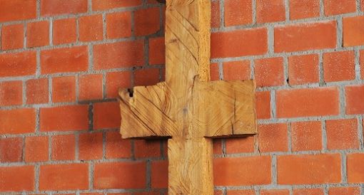 Schmale, symmetrische Holzskulptur vor Backsteinwand, erinnert an Kreuz oder archaisches Zeichen.
