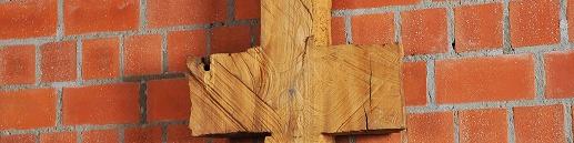 Schmale, symmetrische Holzskulptur vor Backsteinwand, erinnert an Kreuz oder archaisches Zeichen.