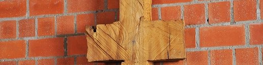 Schmale, symmetrische Holzskulptur vor Backsteinwand, erinnert an Kreuz oder archaisches Zeichen.