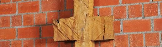 Schmale, symmetrische Holzskulptur vor Backsteinwand, erinnert an Kreuz oder archaisches Zeichen.