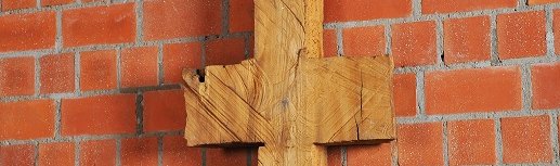 Schmale, symmetrische Holzskulptur vor Backsteinwand, erinnert an Kreuz oder archaisches Zeichen.