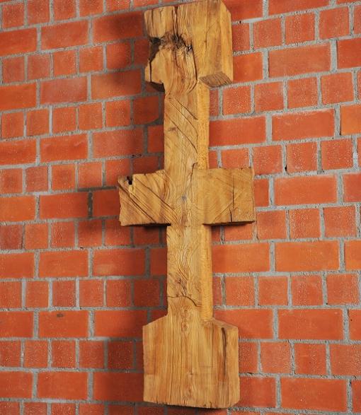 Schmale, symmetrische Holzskulptur vor Backsteinwand, erinnert an Kreuz oder archaisches Zeichen.