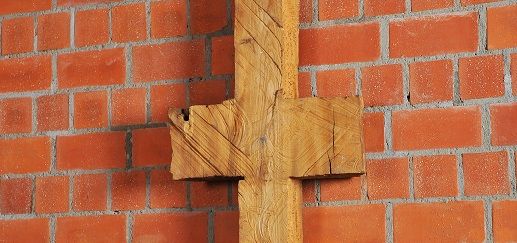 Schmale, symmetrische Holzskulptur vor Backsteinwand, erinnert an Kreuz oder archaisches Zeichen.