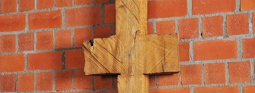 Schmale, symmetrische Holzskulptur vor Backsteinwand, erinnert an Kreuz oder archaisches Zeichen.