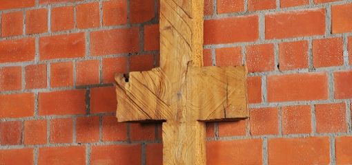 Schmale, symmetrische Holzskulptur vor Backsteinwand, erinnert an Kreuz oder archaisches Zeichen.
