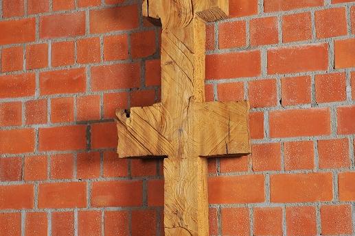 Schmale, symmetrische Holzskulptur vor Backsteinwand, erinnert an Kreuz oder archaisches Zeichen.
