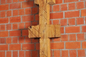 Schmale, symmetrische Holzskulptur vor Backsteinwand, erinnert an Kreuz oder archaisches Zeichen.