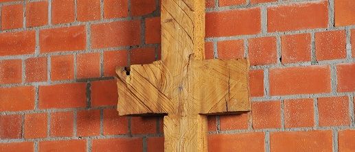 Schmale, symmetrische Holzskulptur vor Backsteinwand, erinnert an Kreuz oder archaisches Zeichen.
