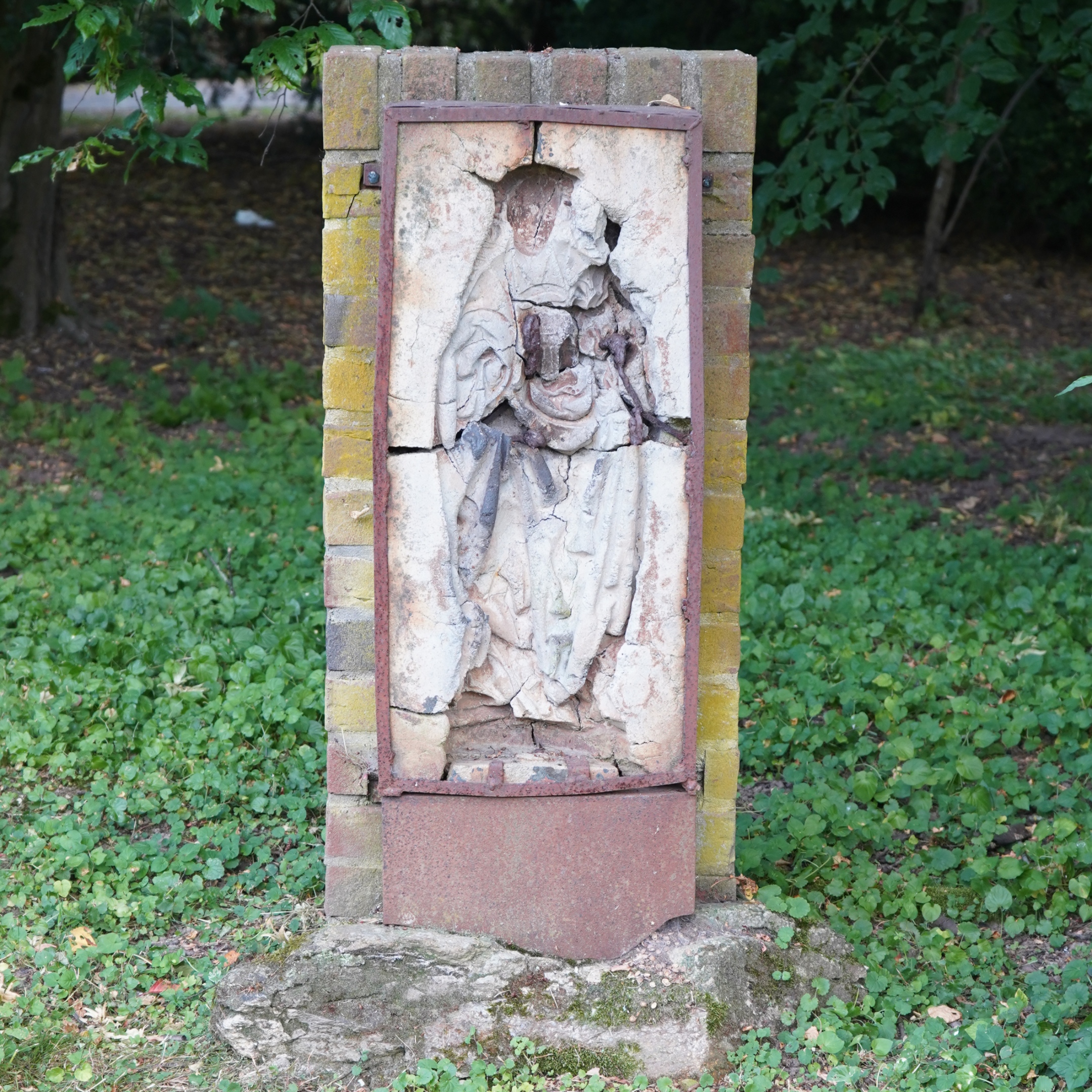 Beschädigte Marienfigur mit fehlendem Gesicht und Kind, im Grünen platziert