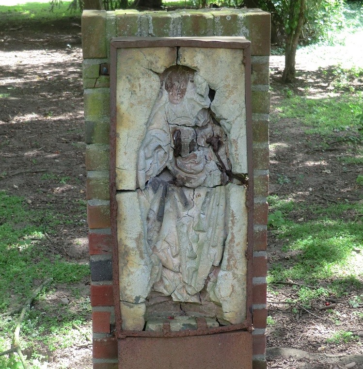 Beschädigte Marienfigur mit fehlendem Gesicht und Kind, im Grünen platziert