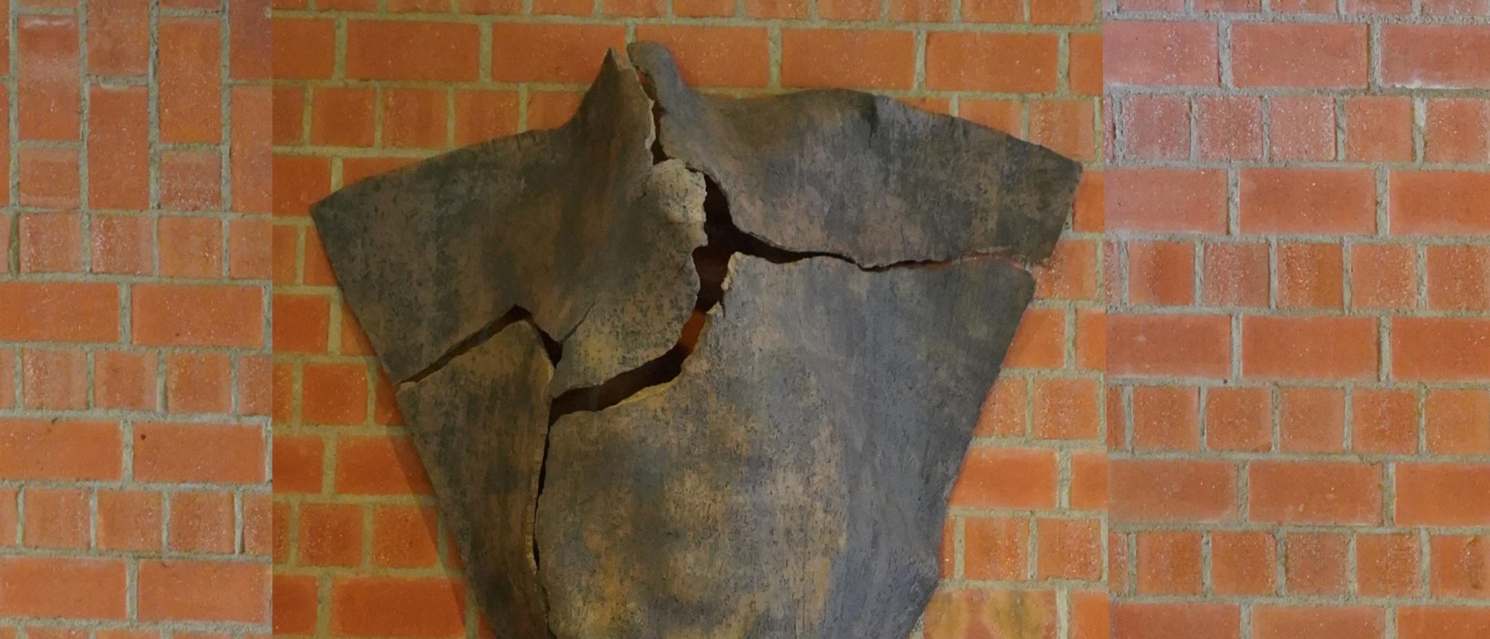 Dreieckige Tonskulptur mit Rissen und angedeuteter menschlicher Silhouette, an der Wand hängend