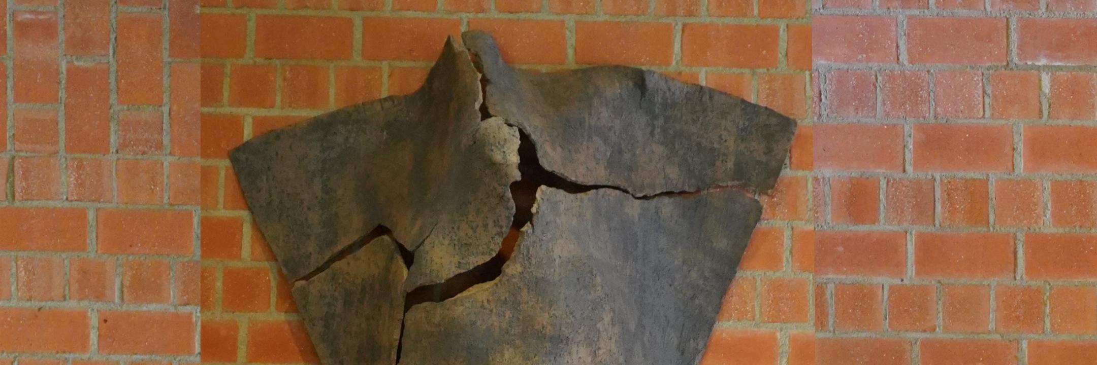 Dreieckige Tonskulptur mit Rissen und angedeuteter menschlicher Silhouette, an der Wand hängend
