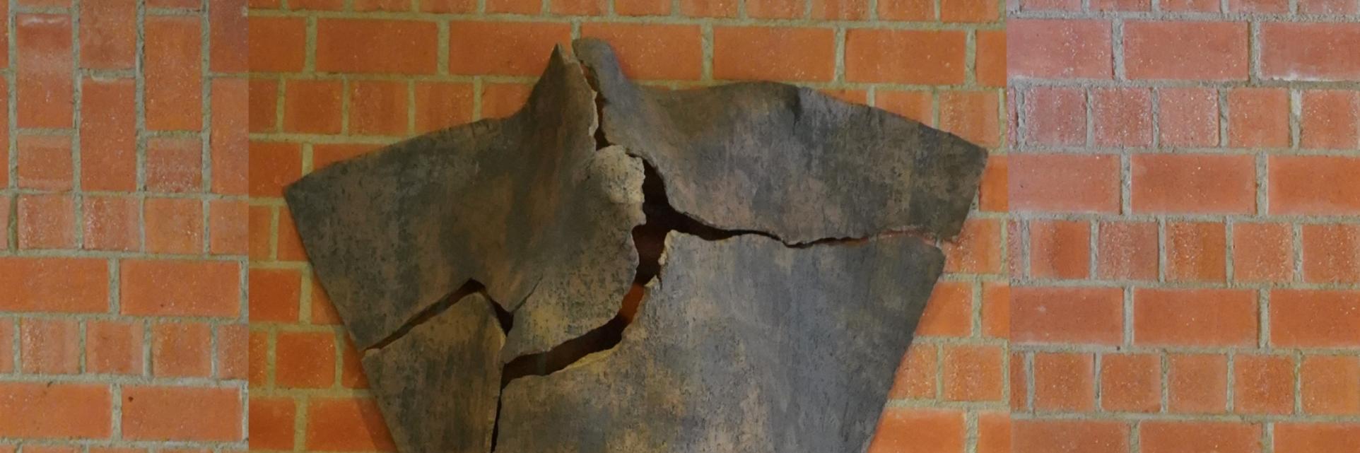 Dreieckige Tonskulptur mit Rissen und angedeuteter menschlicher Silhouette, an der Wand hängend