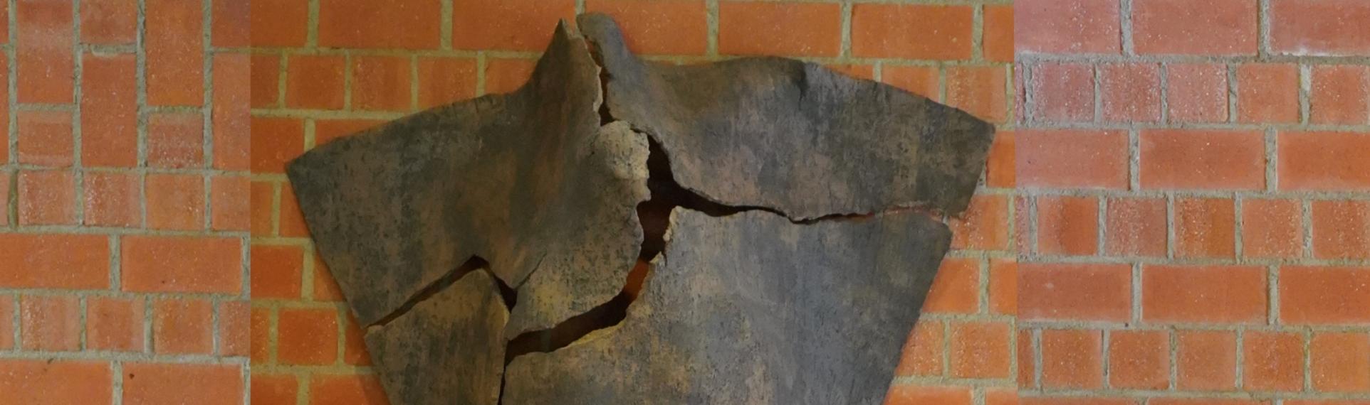 Dreieckige Tonskulptur mit Rissen und angedeuteter menschlicher Silhouette, an der Wand hängend