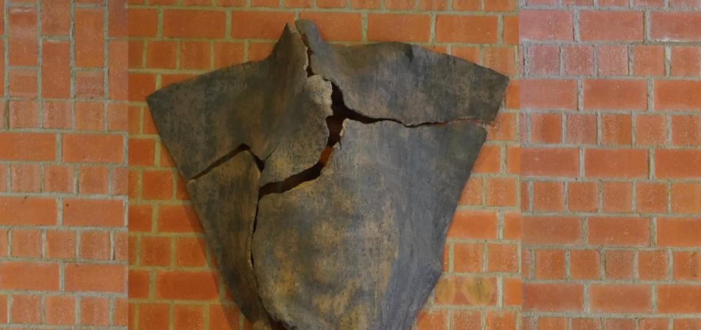 Dreieckige Tonskulptur mit Rissen und angedeuteter menschlicher Silhouette, an der Wand hängend