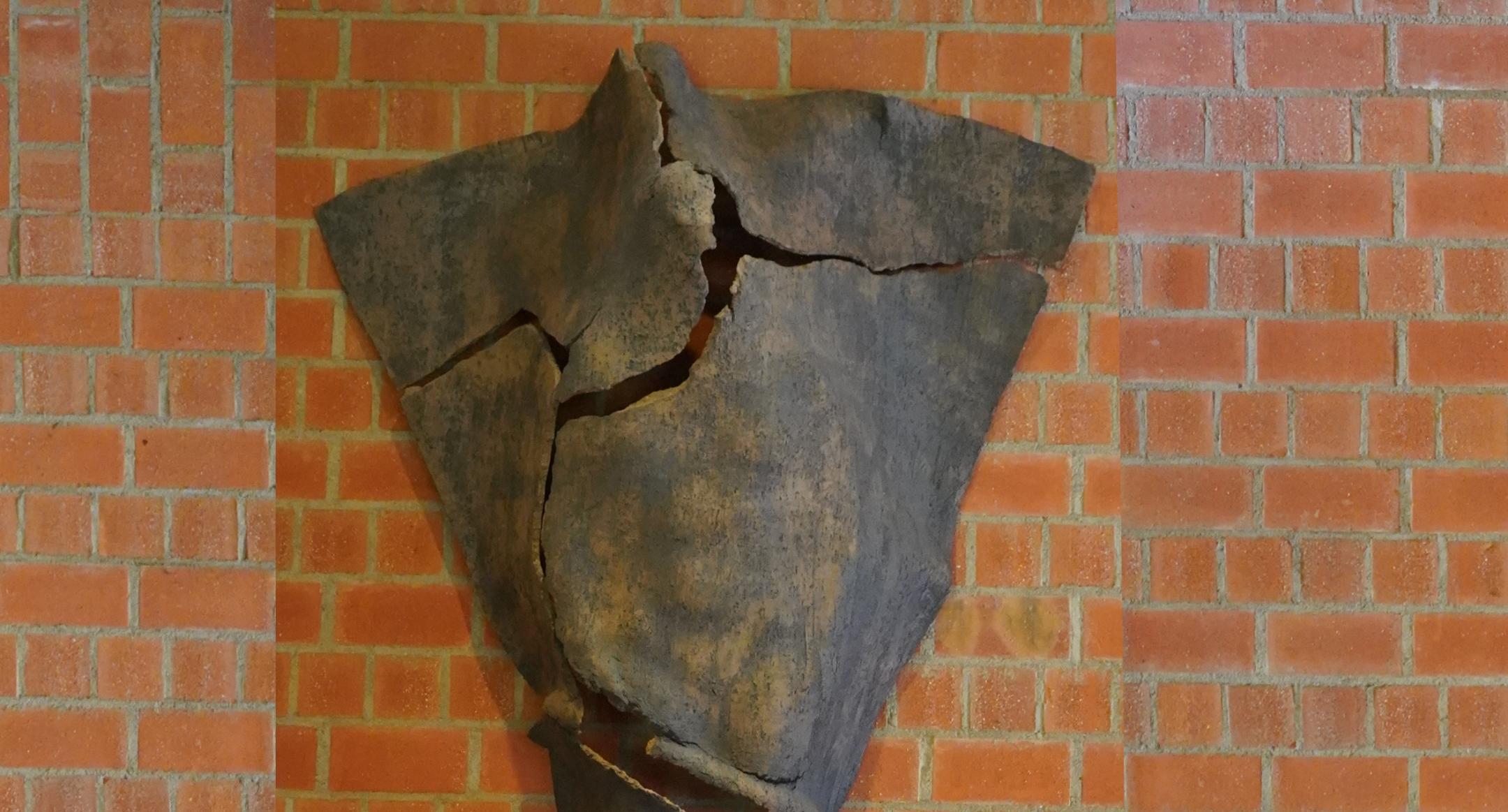 Dreieckige Tonskulptur mit Rissen und angedeuteter menschlicher Silhouette, an der Wand hängend