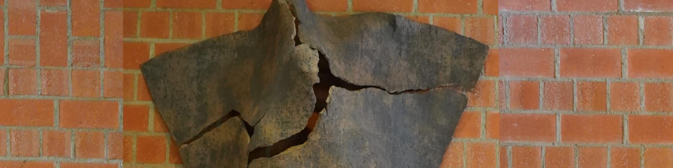 Dreieckige Tonskulptur mit Rissen und angedeuteter menschlicher Silhouette, an der Wand hängend