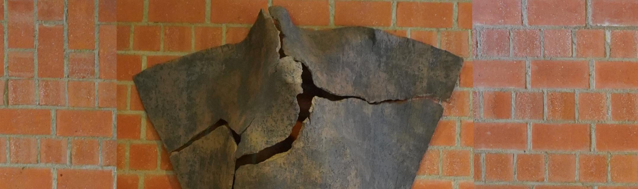 Dreieckige Tonskulptur mit Rissen und angedeuteter menschlicher Silhouette, an der Wand hängend