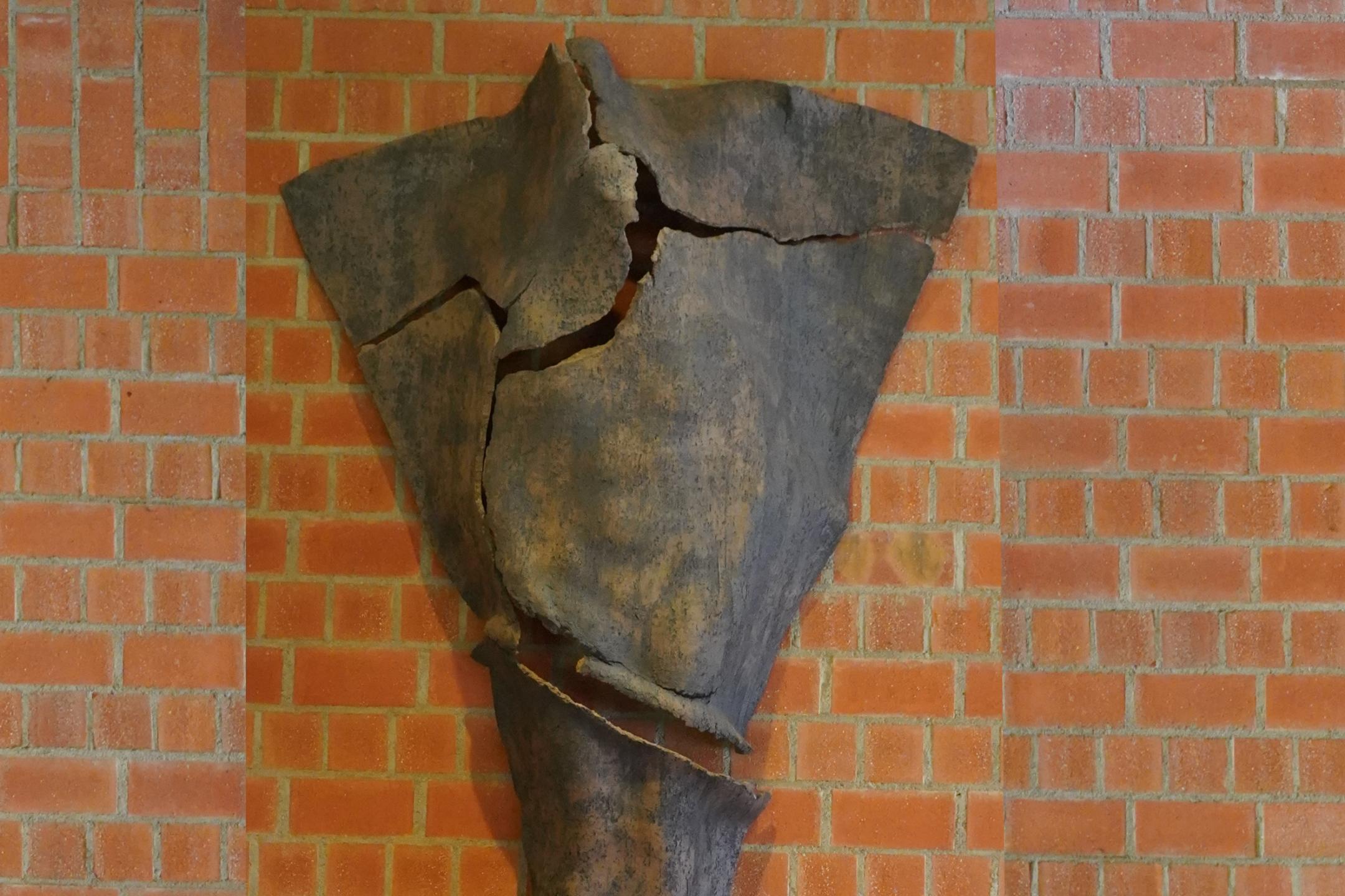 Dreieckige Tonskulptur mit Rissen und angedeuteter menschlicher Silhouette, an der Wand hängend
