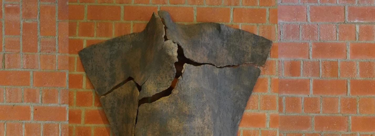 Dreieckige Tonskulptur mit Rissen und angedeuteter menschlicher Silhouette, an der Wand hängend